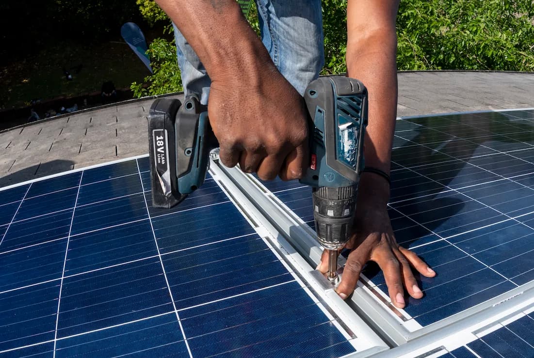 Mains d'un ouvrier utilisant une perceuse pour fixer des panneaux solaires sur un toit.